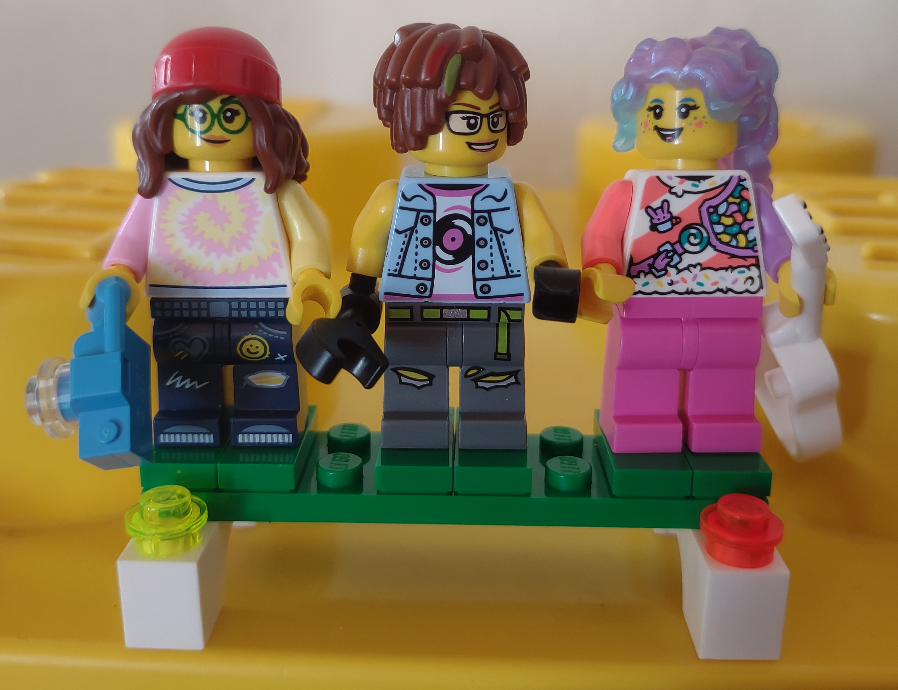 Three LEGO minifigures. The leftmost is wearing a beanie cap, thick round glasses, a tie-dye shirt, and jeans with patches and stitches. She is holding a camera. The middle has shaggy hair with highlights, rectangular glasses, a denim vest, and torn pants. She is holding a wrench. The rightmost has wavy irridescent hair, facepaint, a pink-and white striped shirt with candies on it, and pink pants. She is holding an acousting guitar with gold decoration.